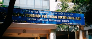 A blue sign with gold letters in both a local language and English, advertising a foreign language specialized school. The sign is situated outside a building surrounded by trees and lit by natural sunlight.