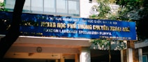 A blue sign with gold letters in both a local language and English, advertising a foreign language specialized school. The sign is situated outside a building surrounded by trees and lit by natural sunlight.