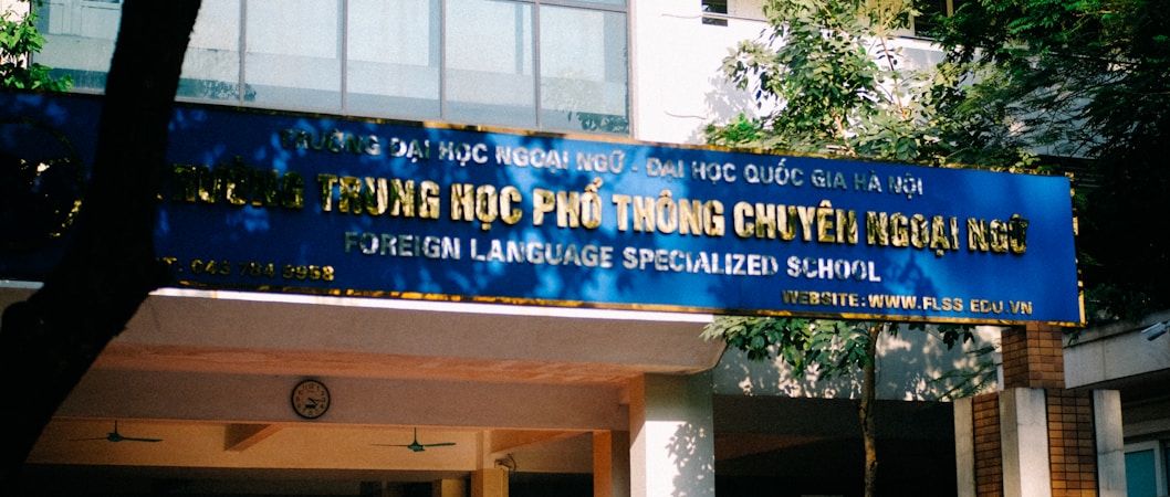A blue sign with gold letters in both a local language and English, advertising a foreign language specialized school. The sign is situated outside a building surrounded by trees and lit by natural sunlight.