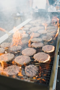 Grilling burgers on an outdoor barbecue grill with visible flames and smoke. Several burger patties are cooking, and a spatula is seen on the left side.