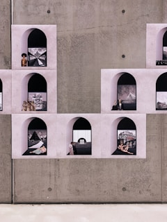 A series of arched wall niches contain various artworks and photographs arranged on a textured concrete wall. Each niche displays a different scene, some featuring classical art images, others modern or abstract scenes. The overall composition creates a unique collage effect.