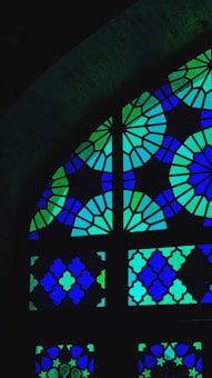 A stained glass window featuring a geometric design with intricate patterns. The glass pieces are arranged in an arch shape, predominantly in shades of blue and green.
