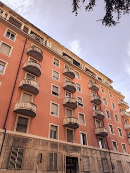 A multi-story residential building with a warm peach-colored facade. Several small balconies with railings are evenly spaced across the building. The structure features numerous windows, some with open shutters, and the bottom portion is lined with stone or concrete. The sky above is clear, and a tree branch partially frames the upper right corner.