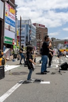 A band performs on a city street, with a lead singer standing at a microphone and a young guitarist playing nearby. The background features tall buildings with colorful advertisements and a bright, partly cloudy sky.