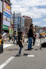 A band performs on a city street, with a lead singer standing at a microphone and a young guitarist playing nearby. The background features tall buildings with colorful advertisements and a bright, partly cloudy sky.