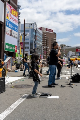 A band performs on a city street, with a lead singer standing at a microphone and a young guitarist playing nearby. The background features tall buildings with colorful advertisements and a bright, partly cloudy sky.