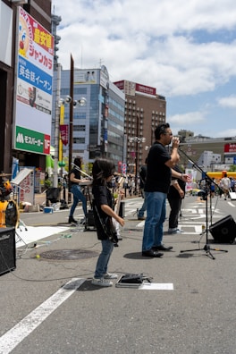 A band performs on a city street, with a lead singer standing at a microphone and a young guitarist playing nearby. The background features tall buildings with colorful advertisements and a bright, partly cloudy sky.