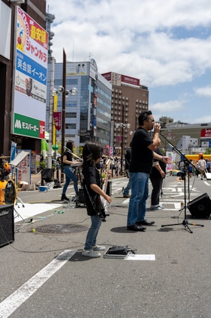 A band performs on a city street, with a lead singer standing at a microphone and a young guitarist playing nearby. The background features tall buildings with colorful advertisements and a bright, partly cloudy sky.
