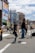 A band performs on a city street, with a lead singer standing at a microphone and a young guitarist playing nearby. The background features tall buildings with colorful advertisements and a bright, partly cloudy sky.