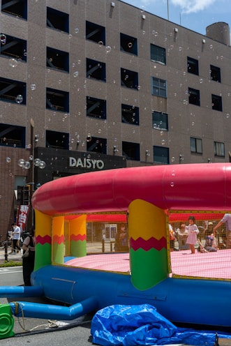 A colorful inflatable bounce house is set up on a city street, surrounded by bubbles floating in the air. Children are playing inside, and a few adults are nearby. The scene is in front of a multi-story building with many windows.