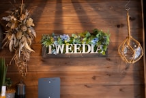 A wooden wall decorated with a floral arrangement and a sign that reads 'TWEEDIA'. The flowers are a mix of dried and fresh-looking blooms, including blue and green hues. To the left, there is a hanging arrangement of dried plants and flowers, and to the right, a rustic hanging glass light fixture with a rope design.
