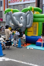 A group of children are gathered around and playing on an inflatable bounce house shaped like an elephant. The bounce house is set up on a city street with safety cones and a rope fence around it. Some adults are assisting the children. Bright colors such as green, blue, and yellow dominate the scene.