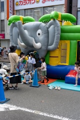 A group of children are gathered around and playing on an inflatable bounce house shaped like an elephant. The bounce house is set up on a city street with safety cones and a rope fence around it. Some adults are assisting the children. Bright colors such as green, blue, and yellow dominate the scene.