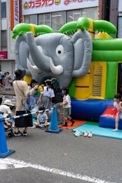 A group of children are gathered around and playing on an inflatable bounce house shaped like an elephant. The bounce house is set up on a city street with safety cones and a rope fence around it. Some adults are assisting the children. Bright colors such as green, blue, and yellow dominate the scene.