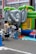 A group of children are gathered around and playing on an inflatable bounce house shaped like an elephant. The bounce house is set up on a city street with safety cones and a rope fence around it. Some adults are assisting the children. Bright colors such as green, blue, and yellow dominate the scene.