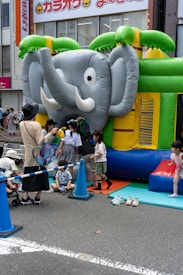 A group of children are gathered around and playing on an inflatable bounce house shaped like an elephant. The bounce house is set up on a city street with safety cones and a rope fence around it. Some adults are assisting the children. Bright colors such as green, blue, and yellow dominate the scene.