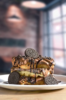 A decadent dessert is served on a white plate, featuring layers of waffles topped with a generous amount of creamy chocolate sauce, drizzles of syrup, and garnished with chocolate sandwich cookies.