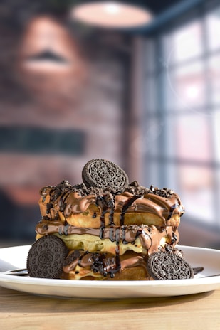 A decadent dessert is served on a white plate, featuring layers of waffles topped with a generous amount of creamy chocolate sauce, drizzles of syrup, and garnished with chocolate sandwich cookies.