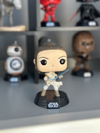 A collection of Star Wars-themed figurines is displayed on a shelf. The main figure in the foreground is a stylized character holding a blue lightsaber. Other figures are blurred in the background, each representing different characters from the franchise.