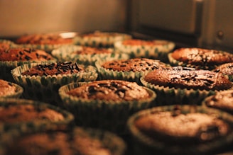 Rows of freshly baked chocolate muffins are nestled in green and white cupcake liners. The muffins appear moist and are topped with sprinkles or chocolate chips. They are arranged closely together inside what appears to be an oven, with a warm glow highlighting their rich texture.