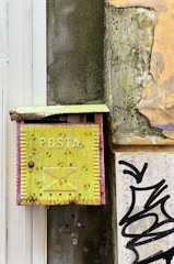 An old, weathered yellow mailbox with 'POSTA' written on it is attached to a dilapidated wall. The mailbox features a hand-painted design with small colorful patterns. The surrounding wall shows signs of decay with peeling paint and exposed concrete. There is also a section of graffiti visible near the bottom right corner of the image.