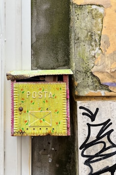 An old, weathered yellow mailbox with 'POSTA' written on it is attached to a dilapidated wall. The mailbox features a hand-painted design with small colorful patterns. The surrounding wall shows signs of decay with peeling paint and exposed concrete. There is also a section of graffiti visible near the bottom right corner of the image.