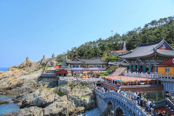 A traditional Asian temple complex nestled on rocky cliffs by the sea, surrounded by lush green trees. The architecture features vibrant colors and ornate designs, with pagoda-style roofs. A crowd of people can be seen walking across a stone bridge leading to the main temple area. Brightly colored lanterns add a festive touch to the scene.