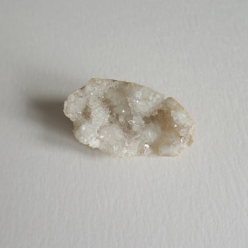A single geode displaying a rough, uneven surface covered in clear and white crystals. The geode is placed on a light, neutral background which enhances its crystalline texture and form.