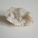 A close-up of a geode with sparkling white crystals on the inside, contrasted against a rough, beige outer shell. The geode rests on a neutral, light-colored surface.