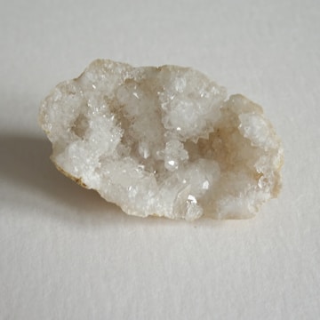 A close-up of a geode with sparkling white crystals on the inside, contrasted against a rough, beige outer shell. The geode rests on a neutral, light-colored surface.