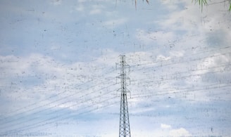 A power transmission tower is reflected in a body of water. The surface of the water is slightly disturbed, creating a shimmering effect. Surrounding the tower, there are power lines stretching across the sky. Some grass or foliage is visible at the top right corner of the image.