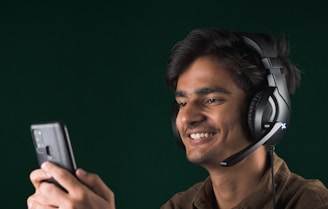 A young man is wearing a headset with a microphone and is looking at a smartphone with a smile. The background is a solid dark color, creating a contrast with his light brown shirt.