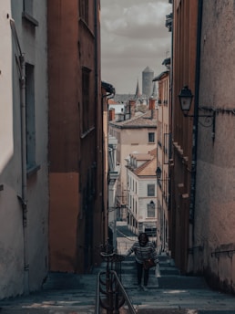 A narrow alleyway flanked by tall buildings leads to an urban cityscape with an iconic tower in the distance. A lone individual ascends the stone staircase, surrounded by warm-toned walls and historical architecture. Street lamps and railings add to the old-world charm.