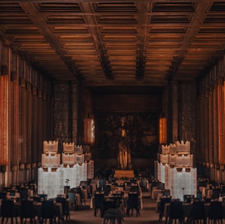 A grand, dimly lit banquet hall with high ceilings and ornate metalwork. Large statues flank a central walkway leading to a prominent statue at the far end. The room is filled with dining tables and chairs, giving an impression of elegance and formality.