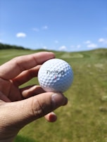 A hand is holding a white golf ball marked with the text 'ProStaff' and the number 1. The background features a lush green golf course under a clear blue sky with a few scattered clouds.