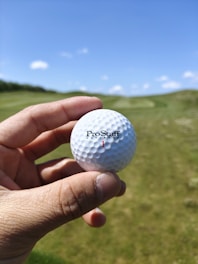 A hand is holding a white golf ball marked with the text 'ProStaff' and the number 1. The background features a lush green golf course under a clear blue sky with a few scattered clouds.