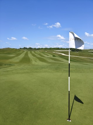 A well-maintained golf course with freshly mowed grass and clear patterns of stripes on the fairway. A white flag on a black and white striped pole is positioned in the putting green, with its shadow visible. The horizon features some bushes and trees under a clear blue sky scattered with a few white clouds.