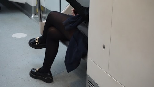 A person sitting on public transportation, dressed in dark clothing with black tights and loafers featuring gold chain details. The interior surroundings are typical of a bus or train, with light-colored walls and gray speckled flooring.