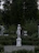 A white classical statue stands gracefully in a garden surrounded by dense greenery. The statue is placed on a pedestal in the center of a paved area, with lush trees in the background and well-maintained bushes and plants around it. An urn-like decorative piece is visible in the background.