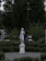 A white classical statue stands gracefully in a garden surrounded by dense greenery. The statue is placed on a pedestal in the center of a paved area, with lush trees in the background and well-maintained bushes and plants around it. An urn-like decorative piece is visible in the background.