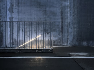 A dimly lit concrete stairwell features a metallic railing and a glowing handrail light. The setting has an industrial feel, characterized by rough textures and a stark, minimalist design.