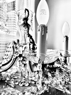 A decorative chandelier with ornate crystal embellishments and candle-shaped bulbs, positioned near a window with a patterned backdrop. The intricate crystals create a visually appealing display of light reflections.