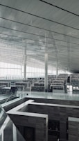 A spacious and modern interior with high ceilings and large windows allowing natural light to flood the space. Multiple levels of bookshelves are visible, creating a sense of a vast library or archive. Minimalist and sleek architectural elements are present, including glass railings and metal beams.