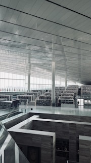 A spacious and modern interior with high ceilings and large windows allowing natural light to flood the space. Multiple levels of bookshelves are visible, creating a sense of a vast library or archive. Minimalist and sleek architectural elements are present, including glass railings and metal beams.