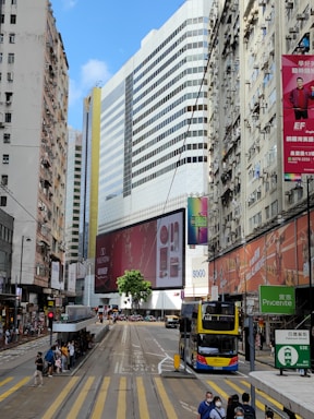 A bustling urban street with tall buildings featuring large advertisements. The scene includes a double-decker bus and people waiting at a bus stop. The architecture is characterized by modern and older high-rise buildings, with one prominently displaying a Valentino ad.