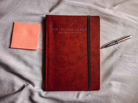 A red notebook with a black elastic strap lies on a light gray fabric surface. To the left of the notebook, there is an orange sticky note, and to the right, there is a metallic pen.