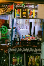 A brightly lit street food stall selling colorful beverages and ice gola. The stall has banners displaying images and names of different flavored drinks such as Rainbow, Mango, Orange, and Kala Khatta. Various bottles of syrups and mixers are neatly arranged on the counter, and disposable cups are hanging on the side. The stall's name, Shahrukh Soda Shikanji Shop Mocktails, is prominently displayed on a banner.