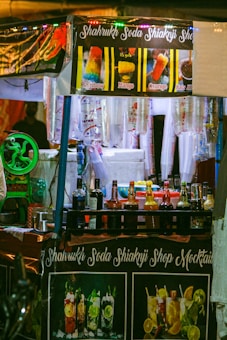 A brightly lit street food stall selling colorful beverages and ice gola. The stall has banners displaying images and names of different flavored drinks such as Rainbow, Mango, Orange, and Kala Khatta. Various bottles of syrups and mixers are neatly arranged on the counter, and disposable cups are hanging on the side. The stall's name, Shahrukh Soda Shikanji Shop Mocktails, is prominently displayed on a banner.