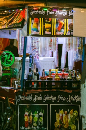 A brightly lit street food stall selling colorful beverages and ice gola. The stall has banners displaying images and names of different flavored drinks such as Rainbow, Mango, Orange, and Kala Khatta. Various bottles of syrups and mixers are neatly arranged on the counter, and disposable cups are hanging on the side. The stall's name, Shahrukh Soda Shikanji Shop Mocktails, is prominently displayed on a banner.