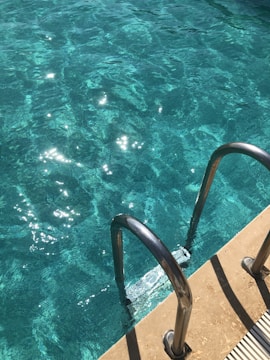 Sunlight reflects off the clear blue water in a swimming pool, highlighting ripples and waves. A metal pool ladder with two handles descends into the water, positioned along the pool edge. The surrounding area is made of a light-colored concrete material.